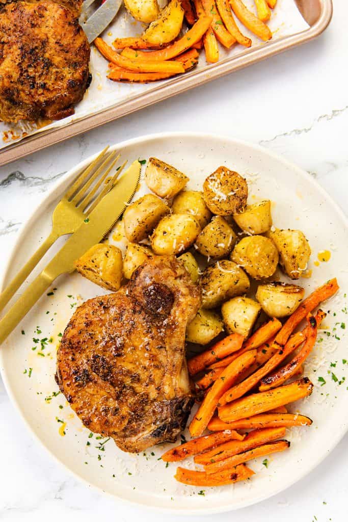 A plate with a seasoned Mediterranean pork chop, roasted potato pieces, and roasted carrot sticks. A fork and knife rest beside the food. Another sheet pan tray with similar food appears above.