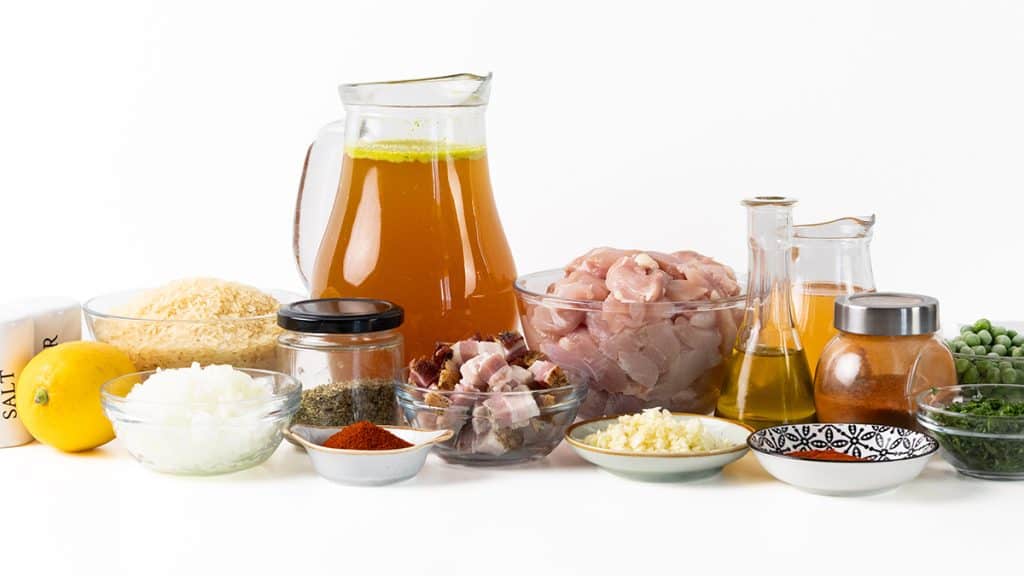 A variety of cooking ingredients for Mediterranean chicken, including chicken, rice, broth, peas, onions, lemon, oil, spices, herbs, and seasonings&mdash;perfectly arranged in bowls and jars on a white surface for your next one-pot meal.
