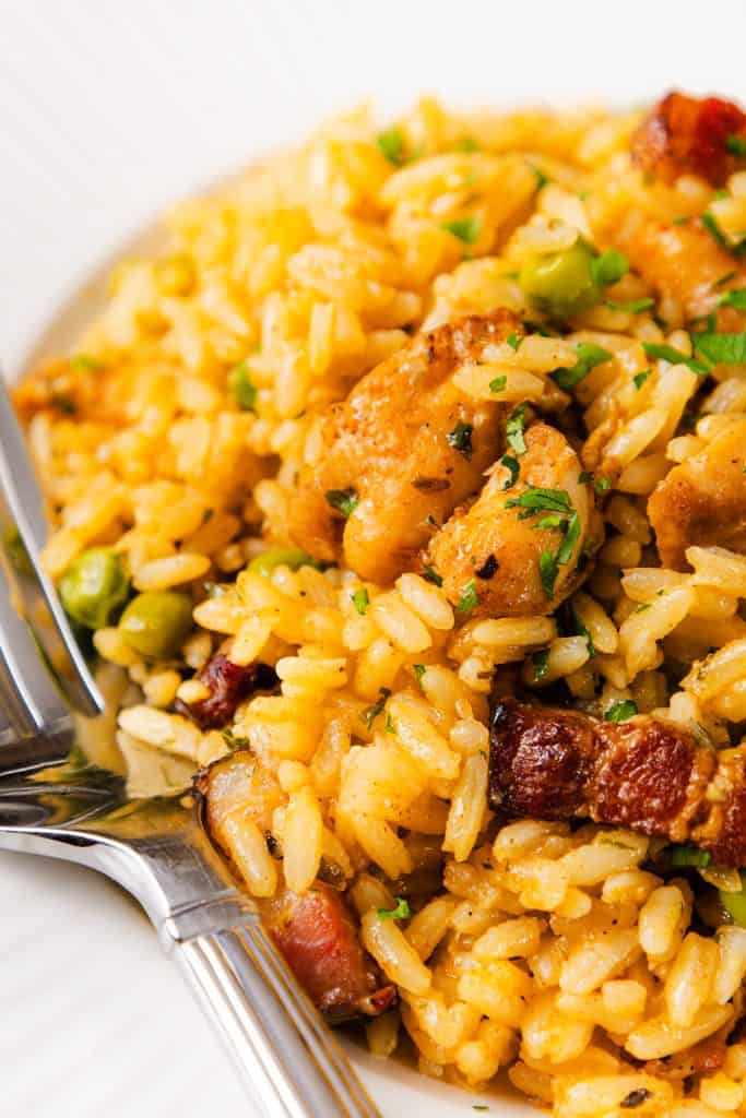 A close-up of a plate of seasoned rice with chunks of Mediterranean chicken, pieces of bacon, green peas, and parsley, served with a fork and spoon.