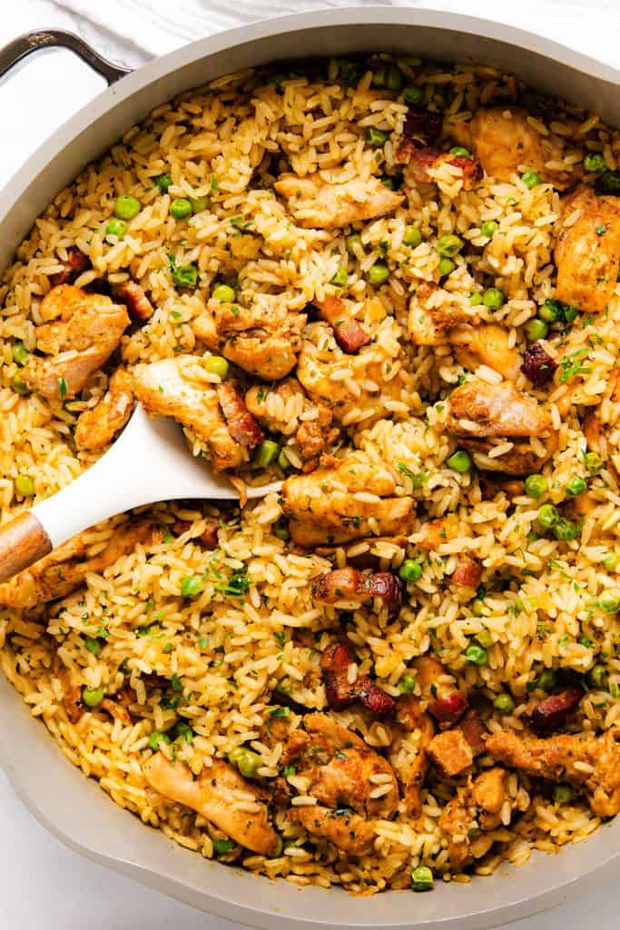 A one-pot pan filled with seasoned rice, pieces of Mediterranean chicken, bits of bacon, and green peas. A white spoon rests in the rice, ready for serving.
