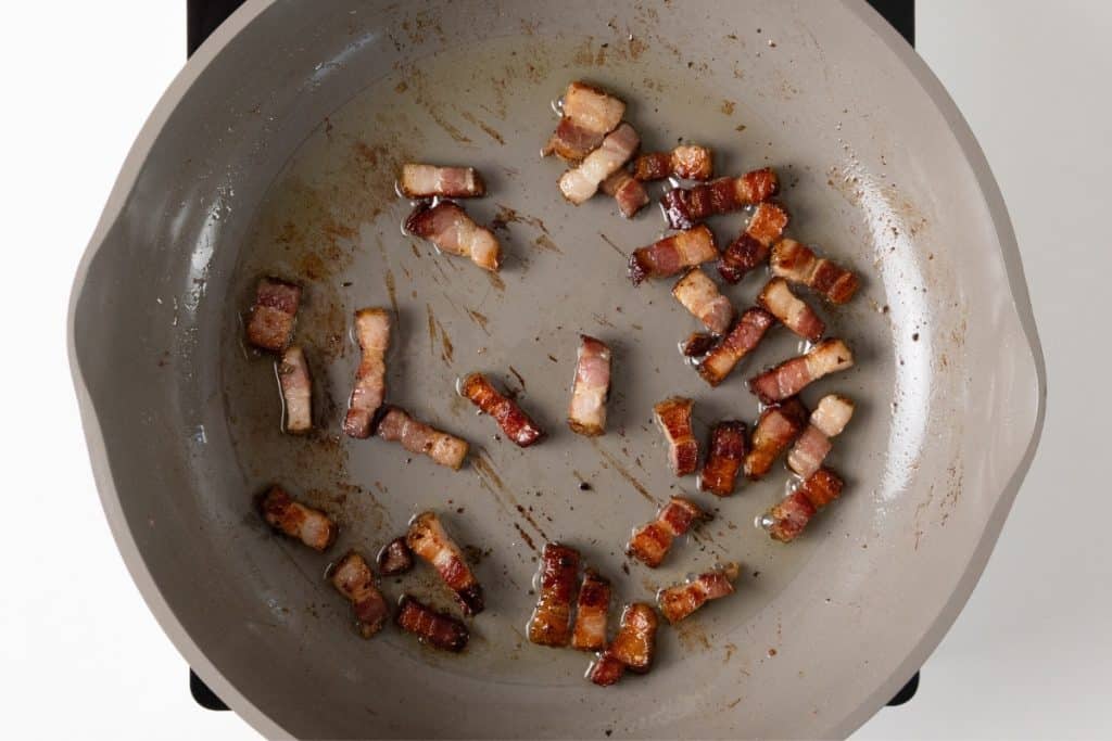 Small pieces of crispy bacon are scattered in a light gray frying pan, with some oil and brown bits visible on the pan&rsquo;s surface&mdash;perfect for starting a flavorful one-pot chicken or Mediterranean chicken recipe.