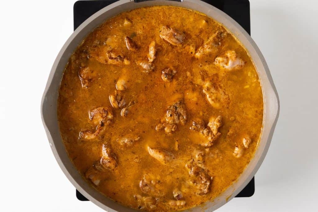 A pot filled with chicken pieces simmering in a rich, orange-brown sauce on a black stovetop, viewed from above&mdash;this one-pot Mediterranean chicken is the perfect blend of vibrant flavors and comforting simplicity.