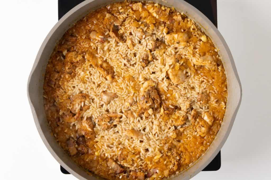 A pan filled with cooked rice and chunks of Mediterranean chicken in a rich, orange-colored sauce, shown from above on a white and black background&mdash;an inviting one-pot recipe perfect for easy, flavorful meals.