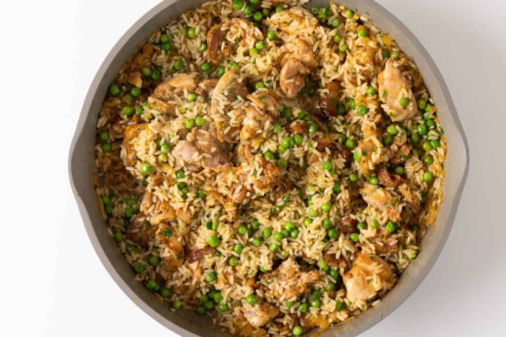 A skillet filled with cooked rice, pieces of chicken, green peas, and small bits of herbs, all mixed together in a savory, golden-brown one-pot chicken and rice dish.