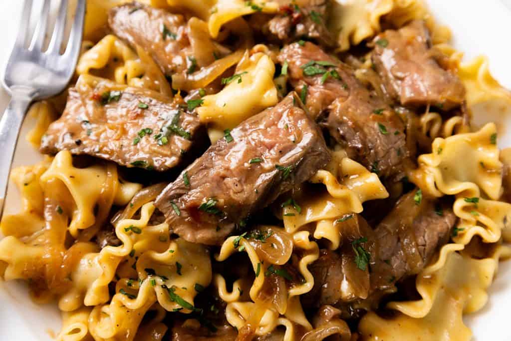 A plate of ruffled pasta tossed with steak strips in a sherry onion gravy, garnished with herbs, with a fork on the left.