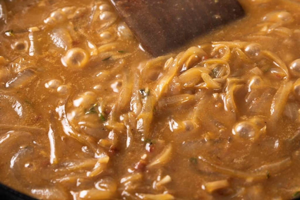 Onions caramelizing in a bubbling brown sauce in a pan with a wooden spoon.