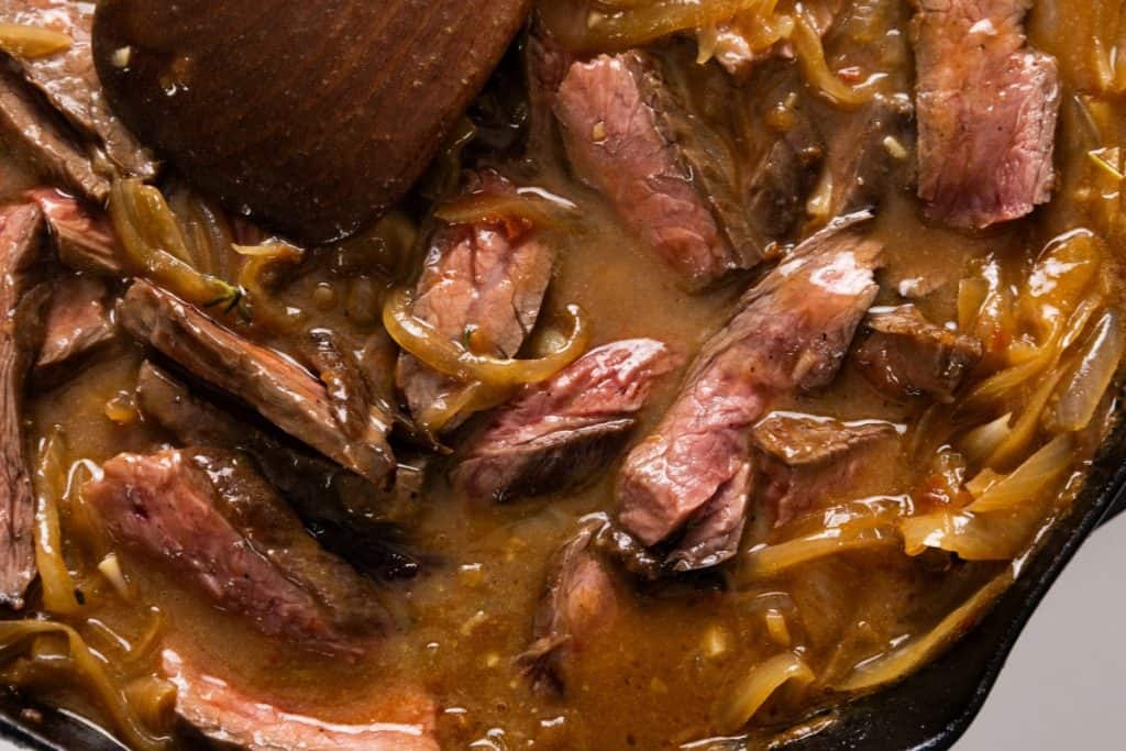 Sliced steak with caramelized onions in a savory brown sauce being stirred with a wooden spoon in a skillet.