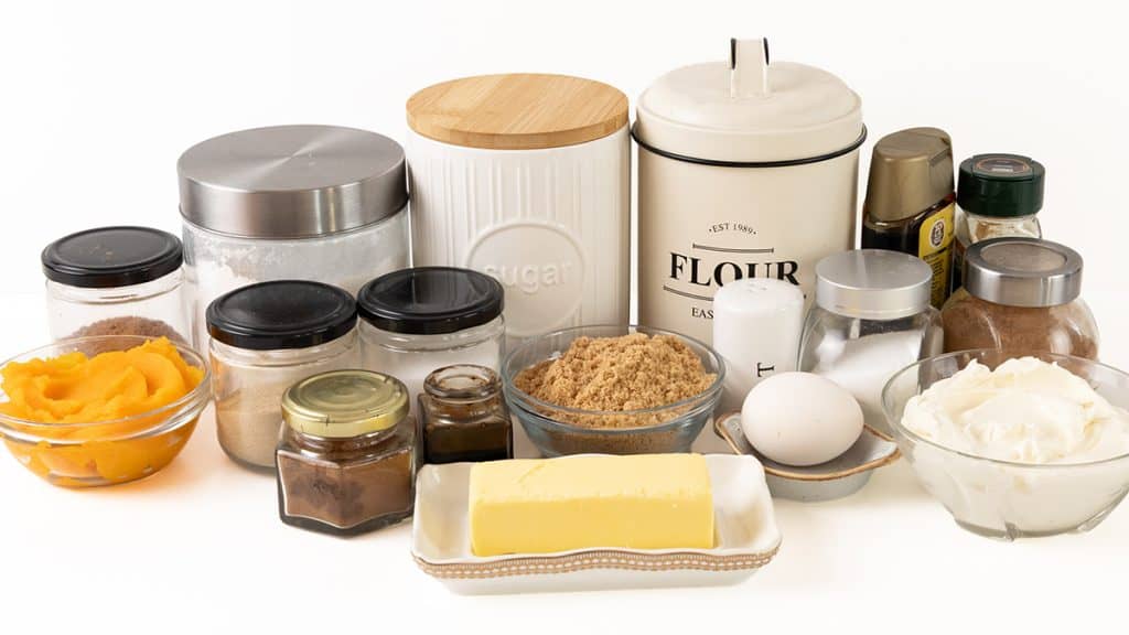 An assortment of baking ingredients for pumpkin spice cookie bars including bowls of pumpkin puree, brown sugar, and cream cheese, a stick of butter, an egg, and several jars and canisters of flour and sugar.