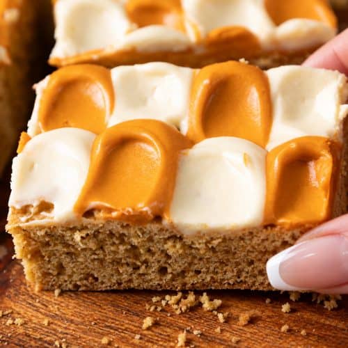 A hand picks up a square pumpkin spice cookie bar topped with a checkerboard pattern of white and caramel-colored frosting.