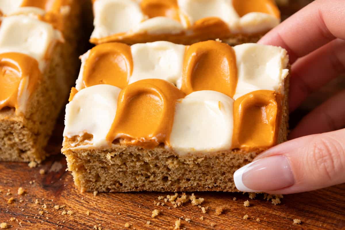 A hand picks up a square pumpkin spice cookie bar topped with a checkerboard pattern of white and caramel-colored frosting.