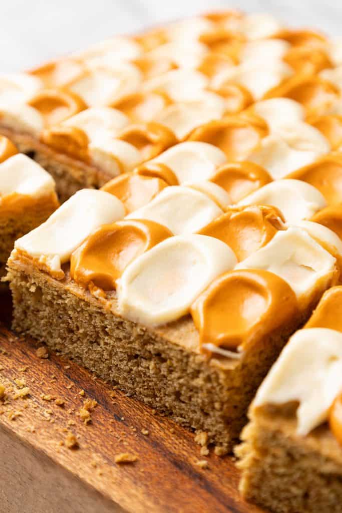 A square of pumpkin spice cake topped with alternating dollops of white and caramel-colored frosting on a wooden board.