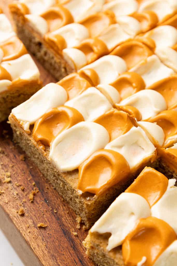 Square slices of pumpkin spice sheet cake topped with a checkerboard pattern of white and caramel-colored frosting on a wooden board.