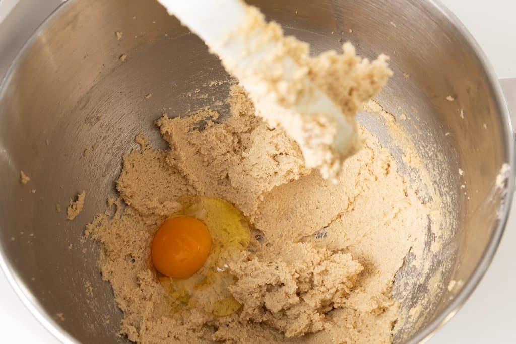 A cracked egg sits in a metal mixing bowl containing creamed butter and sugar for pumpkin spice cookie bars, with a spatula lifting a portion of the mixture.