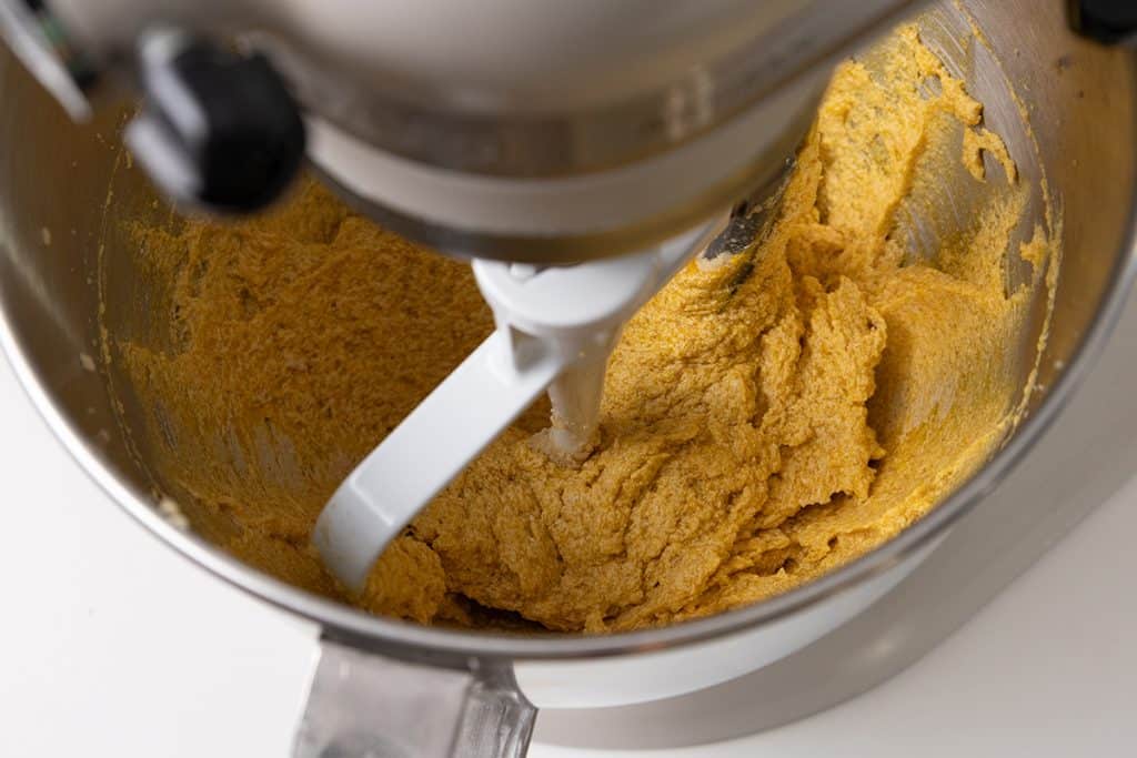 Stand mixer mixing thick yellow dough for pumpkin spice cookies in a metal bowl.